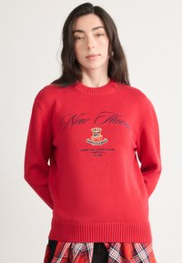 Rød strikket sweater med ribbet udskæring og manchetter. Har broderet tekst og logo i blå, parret med et ternet nederdel.