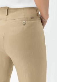 Pantaloni in lino beige con un design aderente, tasca posteriore con bottone e una texture liscia. La fascia della vita presenta un dettaglio logo discreto.