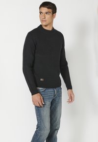 Strickpullover in dunklem Marineblau mit strukturiertem Muster, rundem Halsausschnitt und gerippten Bündchen. Getragen zu blauen Jeans, beinhaltet ein kleines Logo-Patch.
