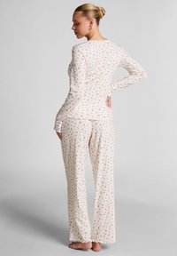 Frau steht barfuß in einem weißen Pyjama-Set mit kleinem rosa Blumenmuster, langärmlig und mit weitem Bein, mit dem Rücken zur Kamera und der Hand auf der Hüfte.