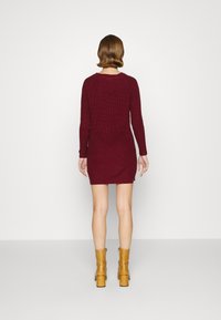 Abito a maglia bordeaux con maniche lunghe e texture a costine, caratterizzato da una silhouette aderente. Abbinato a stivali alla caviglia gialli su uno sfondo semplice.
