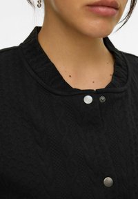 Giacca nera texturizzata con collo a coste, bottoni a scatto e un motivo lavorato a maglia con dettagli a rombi e a treccia.