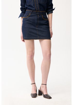 Frau trägt dunkelblaue Jeans-Minirock mit orangefarbenen Nähten, passende Jeansbluse und schwarze Blockabsatz-Sandaletten mit Knöchelriemen vor weißem Hintergrund.