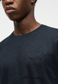 Νεαρό άτομο που φοράει μπλε σκουρό μπλουζάκι με στρογγυλή λαιμόκοψη και μικρή τσέπη στο στήθος, εμφανίζεται από τον λαιμό έως το πάνω μέρος του στήθους, μπροστά σε απλό φόντο.