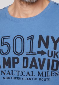 Blaues Baumwoll-T-Shirt mit schwarzem Text, der Zahlen und Wörter wie "501", "NY", "UK" und "CAMP DAVID" zeigt, in einem Rundhalsausschnitt.