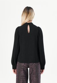 Kvinne med langt blondt hår har på seg en svart langermet bluse med en nøkkelhullåpning bak og bukser med lilla leopardmønster, stående mot hvit bakgrunn.