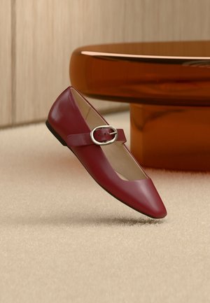 Chaussure plate Mary Jane en cuir rouge avec une bride à boucle argentée, posée sur un tapis beige près d'une table ronde marron.