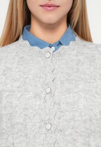 Lys grå, strikket cardigan med kabelmønster og bølget udskæring, der har tre knaplukninger og en blå denimskjorte under.