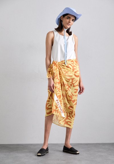 Wrapped skirt in yellow with orange coral pattern, paired with a white tank top. Accompanied by a light blue hat and black espadrilles.