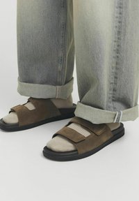 Sandali in suede marrone con suola nera, dotati di due cinghie regolabili. Indossati con jeans grigi chiari, leggermente larghi e arrotolati.