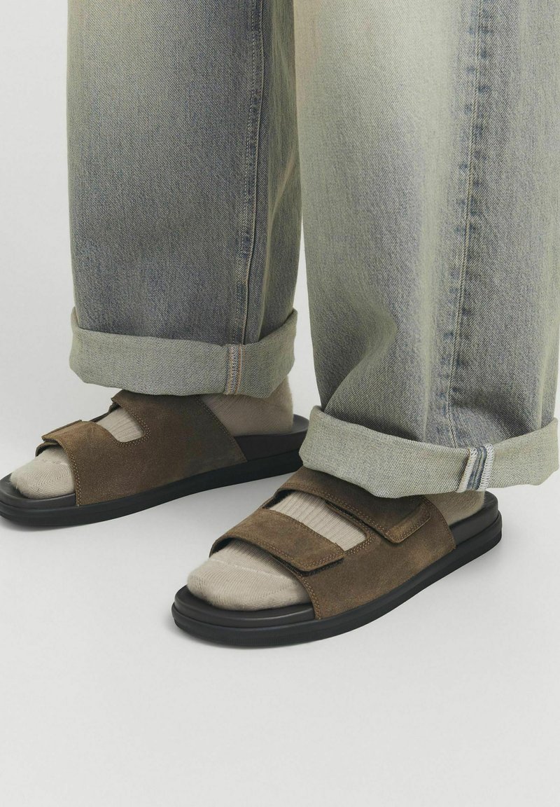 Sandali in suede marrone con suola nera, dotati di due cinghie regolabili. Indossati con jeans grigi chiari, leggermente larghi e arrotolati.