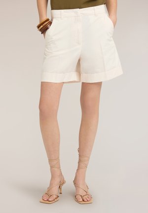 Femme portant un short ajusté écru avec revers, un haut vert olive, des bracelets en bois et des sandales à talons beiges à lacets.