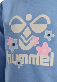 Sweatshirt bleu clair avec un grand motif beige orné de designs floraux en rose, bleu clair et noir, et le texte "hummel".