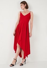Rotes ärmelloses Kleid mit V-Ausschnitt, gerafftem Oberteil und asymmetrischem Saum. Verfügt über einen fließenden Rock und schwarze High-Heels.