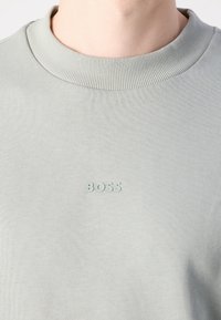 Gros plan d'un sweat-shirt gris clair côtelé à col rond avec le mot « BOSS » imprimé en petites lettres en relief sur la poitrine.