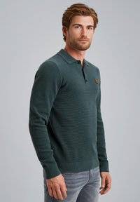Polo de manga larga verde con tejido texturizado, tapeta de botones y un pequeño logo en el pecho. Combinado con unos jeans gris claro.