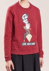 Sudadera de algodón roja con mangas largas, cuello y puños acanalados, que presenta un gráfico de un personaje estilizado y el texto "Love Moschino."