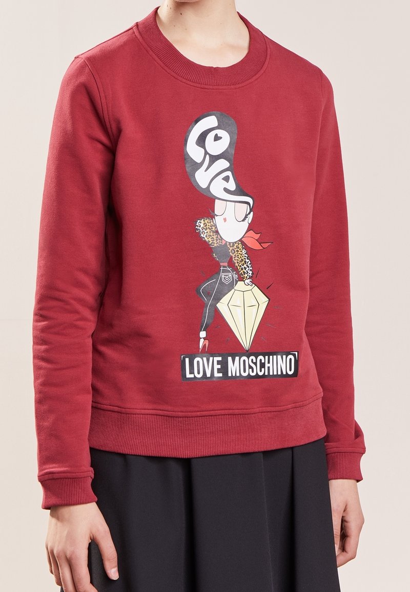 Sudadera de algodón roja con mangas largas, cuello y puños acanalados, que presenta un gráfico de un personaje estilizado y el texto "Love Moschino."