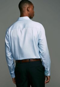 Hellblaues Langarmhemd mit Button-down-Kragen, sichtbaren Nähten und glatter Textur, getragen mit dunklen Hosen und einem braunen Gürtel.