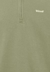 Jersey verde con cuello con cremallera, hecho de una tela suave. Presenta un pequeño logo "blend" en blanco en el pecho. Textura suave, diseño informal.