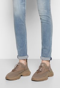 Scarpe da ginnastica marrone chiaro in materiale a rete, superficie texturizzata, punta arrotondata e design unico della suola. Indossate con jeans azzurri a maniche risvoltate.