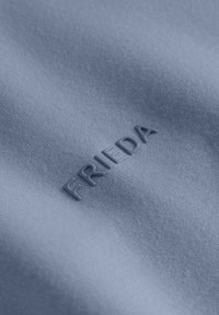 Strukturalny, jasnoniebieski materiał z wytłaczanym logo "FRIEDA", podkreślający gładką powierzchnię z delikatnymi różnicami w odcieniu.