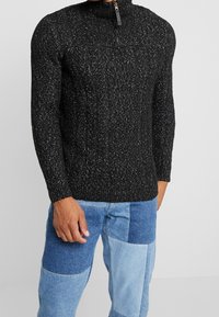 Pull en tricot noir avec un col montant et une fermeture éclair, présentant un motif texturé. Porté avec un jean patchwork bleu clair.