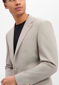 Mann trägt eine hellbeige Anzugjacke über einem schwarzen Hemd, das auf Taillenhöhe zugeknöpft ist, mit neutralem Gesichtsausdruck und schaut zur Seite.