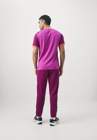 Maglietta sportiva a maniche corte di colore rosa scuro con righe arancioni, abbinata a pantaloni rosa scuro, indossata con sneakers bianche e nere.
