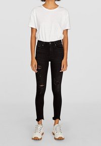 Jeans skinny negros de talle alto con detalles desgastados, combinados con una camisa blanca de manga corta con acentos brillantes y zapatillas blancas.