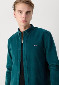 Camisa de botones de sarga en color azul verdoso con cuello, que presenta un pequeño logotipo bordado en el pecho y una superficie texturizada.