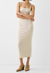 Beige, figursydd klänning i knälängd med fyrkantig halsringning och tunna remmar. Tillverkad av ett glatt, elastiskt tyg. Bärs med beige träskor.