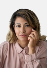 Kvinne med lysebrunt, bølgete hår, iført en lys rosa skjorte med krage. Hun berører haken med en hånd som viser en delikat ring.