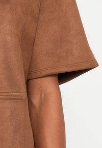 Bruin suède kraagloos kledingstuk met korte mouwen op een persoon, met een zichtbaar gladde arm en duidelijke stikdetails op de stof.