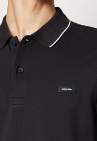 Polo noir avec deux boutons, bordure blanche sur le col, et petit logo rectangulaire Calvin Klein sur la poitrine gauche.