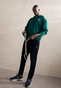 Zöld cipzáras kapucnis pulóver logóval, fekete nadrág és tengerészkék sportcipő. Egy bézs kötelet tart a kezében. Simára szabott anyagok modern, sportos fazonnal.