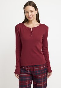 Camisa de manga larga color burdeos con cuello tipo henley y tres botones, hecha de una tela texturizada, combinada con pantalones de pijama a cuadros en rojo y azul marino.