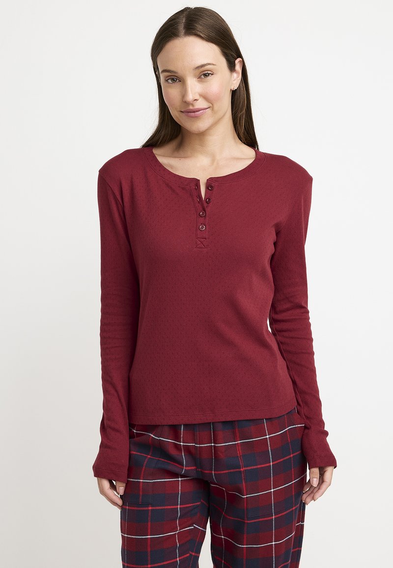 Camisa de manga larga color burdeos con cuello tipo henley y tres botones, hecha de una tela texturizada, combinada con pantalones de pijama a cuadros en rojo y azul marino.