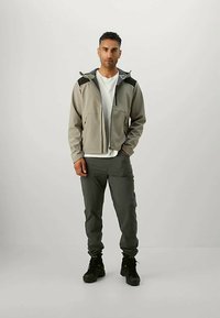 Lysegrå fleecejakke med sorte detaljer, lynlåslukning, to frontlommer, parret med mørkegrå bukser og sorte støvler. Modellen står oprejst.