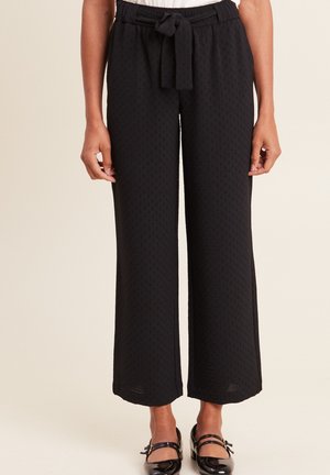 Pantalons larges noirs avec un motif texturé, dotés d'une taille élastique et d'une ceinture en tissu. Des ballerines noires brillantes complètent le look.