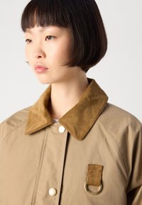 Tan jasje met een corduroy kraag, gouden accenten en een patch met een ring. Glad textuur met een minimalistisch design.