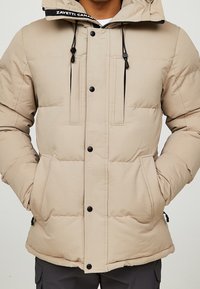 Chaqueta de plumas beige con capucha, con cordones negros, dos bolsillos frontales y botones a presión. Textura suave con diseño acolchado.