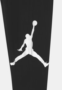 Schwarze Hose mit einer großen weißen Silhouette einer springenden Figur mit einem Basketball, aus weichem Stoff, minimalistischem Design.