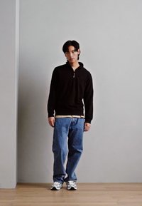 Sort zip-up trøje over en lys skjorte, parret med lyseblå jeans og hvide sneakers. Glat stof, standard pasform, afslappet design.