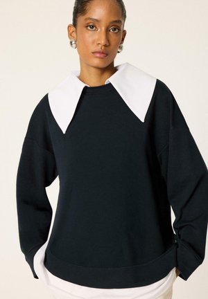 Femme portant un haut noir à manches longues avec un col blanc pointu oversize et de grandes boucles d’oreilles rondes argentées.