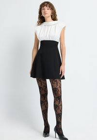 Vrouw in een zwarte A-lijn rok, een witte mouwloze blouse, patronen zwarte doorschijnende panty's en zwarte hoge hakken, staande op een witte achtergrond.