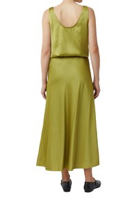 Vrouw in een mouwloze olijfgroene satijnen midi-jurk met een ronde rug en zwarte studs flats, staand met de rug naar de kijker gericht.