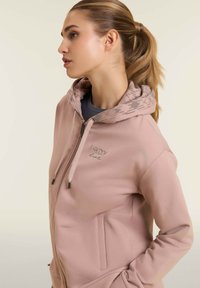 Felpa con zip di colore rosa chiaro, con cappuccio decorato, presenta un logo ricamato sul petto e tasche laterali. Tessuto morbido.