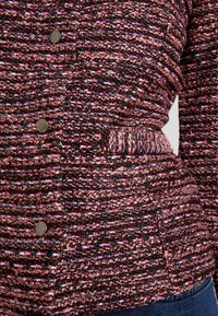 Gebreid jack in donkerbruin, roze en marineblauw. Heeft een gestructureerde stof, metalen knopen en een zijvak voor extra functionaliteit.