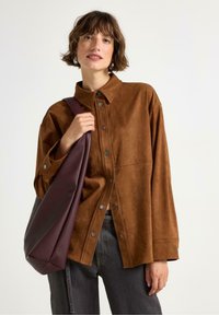 Camicia in suede sintetico marrone con bottoni a pressione, vestibilità oversize, maniche lunghe, abbinata a una borsa slouchy di un marrone scuro, indossata su uno sfondo neutro.